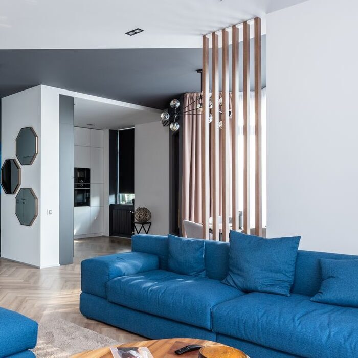 Interior of stylish flat with blue sofa and pouf near wooden table placed near kitchen with white cabinets and decorations on wall