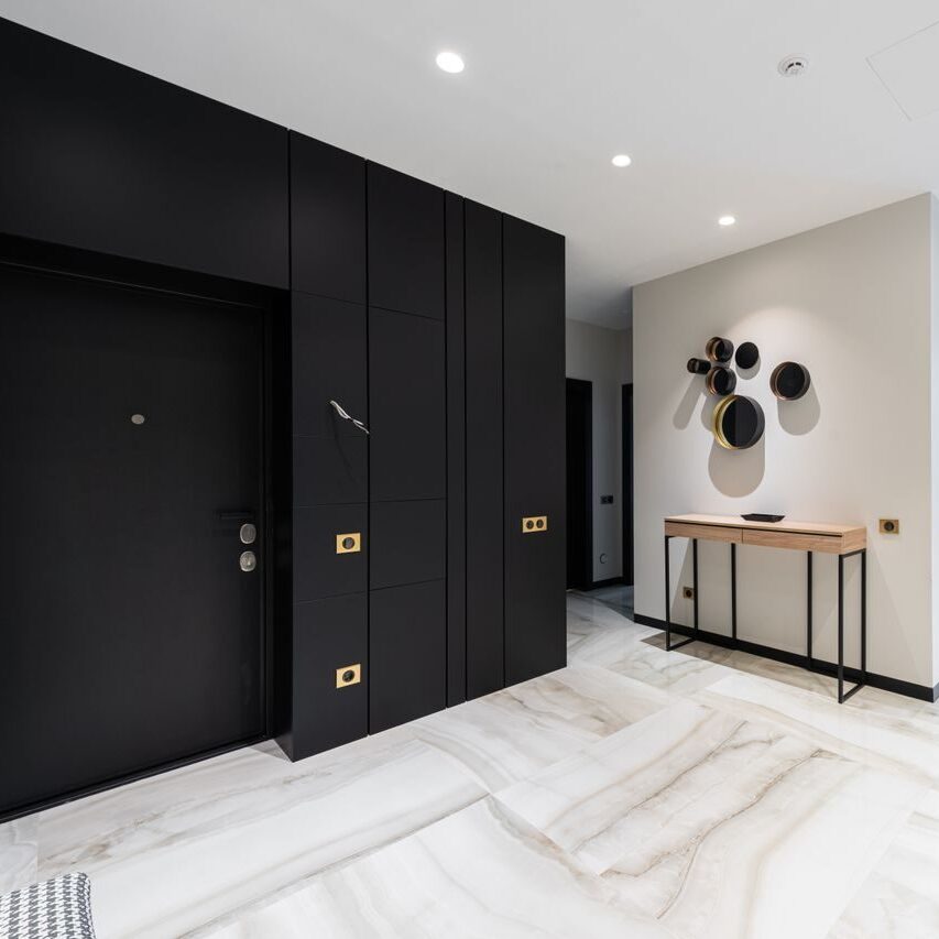 Interior of modern light hall with black doorway and stylish table at wall with entrance to open plan kitchen with black island conuter